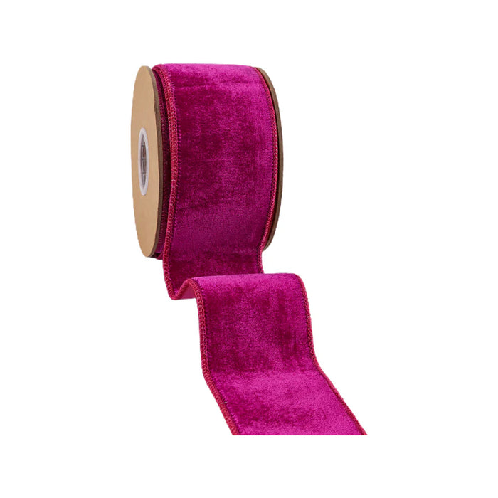 A spool of 2.5-inch Hot Pink Premium Velvet Ribbon with Pink Dupioni Backing (7049W-185-63C) is partially unrolled, showcasing its vibrant hue. The glossy texture and stitched edges add to the ribbon's elegance as it is wrapped around a cardboard core and stands upright against a plain white background.