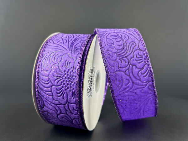 Two rolls of the 1.5"X10Y Purple Glittered Flower Embossed Ribbon 72433-09-11 are displayed against a dark background. The partially unwrapped ribbons showcase their intricate floral designs, while the wired edges ensure they maintain shape, adding elegance and flair to any decorative project.