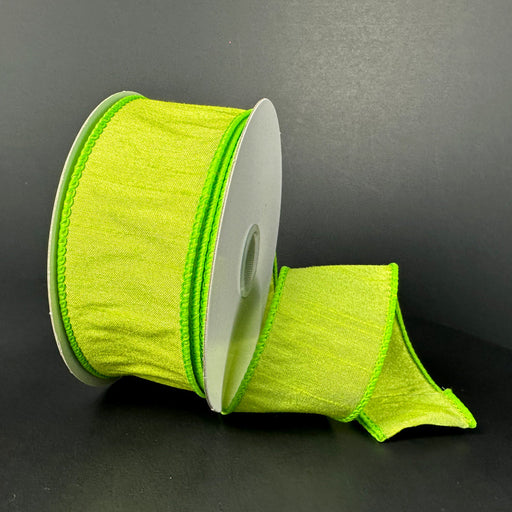 A spool of 1.5X10Y Lime Faux Dupioni Ribbon (72434-09-09), partially unrolled, rests on its side against a dark background, showcasing its textured look and stitched edges.