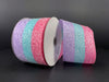 Two rolls of the 2.5"X10Y Pink-Lt Blue-Pearl Candied Striped wired ribbon (product code: 76358-40-44) are showcased against a dark backdrop. Each ribbon features horizontal stripes in purple, teal, and pink-light blue-pearl colors, and they possess a sparkling texture that beautifully catches the light.