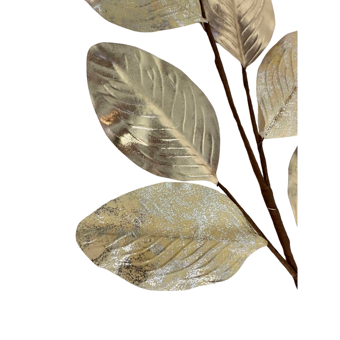 The 27" Champagne Metallic Magnolia Leaves Spray ISB78100 showcases metallic magnolia leaves with a champagne sheen. The leaves have a shiny, textured surface and are elegantly attached to a brown stem set against a white background.