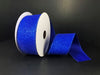 A spool of 1.5"X10Y Royal Blue All Flat Glitter Ribbon X820609-25 glistens on a black surface. Partially unrolled, it reveals its vibrant shimmer and rich color. The visible ends of the ribbon highlight its intricate texture and wired edges, making it perfect for crafting elegant decorations.