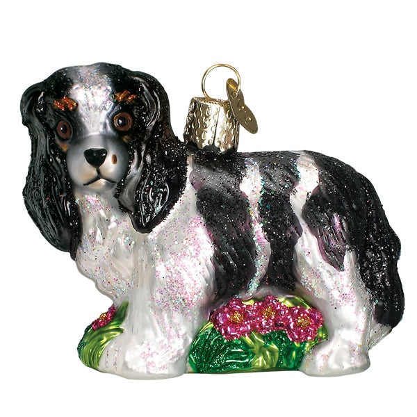 The Black/White King Charles Spaniel Ornament by Old World Christmas (product number 12612) features a black and white King Charles Spaniel with long ears, embellished with glitter. This representation of man's best friend stands on a green base adorned with pink flowers, topped off with a gold cap and loop for easy hanging.