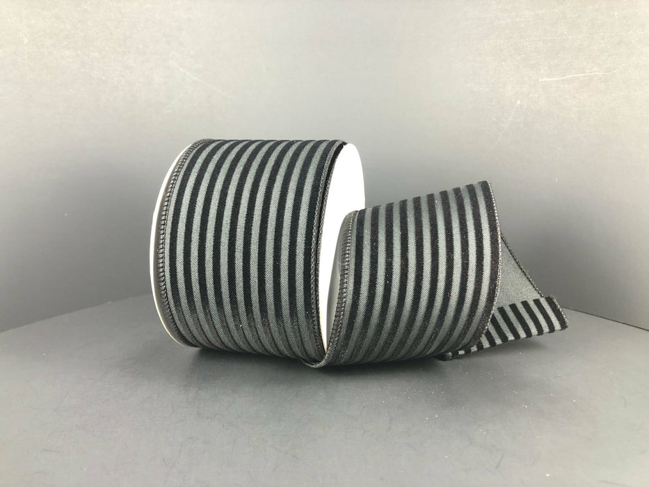 A spool of Black/Black Flocked Thin Cabana Stripes, 2.5X10Y Ribbon 71209-40-21 rests on a gray background, partially unrolled to showcase its smooth matte texture and elegant wired edges.