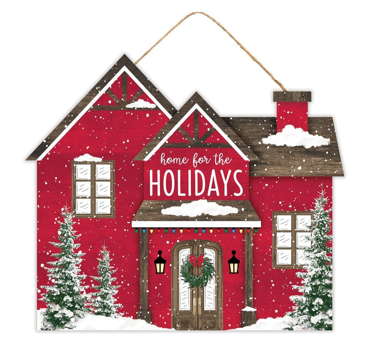 An enchanting 11.25"L x 9.75"H MDF house sign, named the Home/Holidays House (AP7174), captures the spirit of a cozy home with its red structure, brown and white snow-covered roofs, and inviting "Home For The Holidays" message. It features a festive wreath on the door, vibrant lights, delicate snowflakes, and is framed by two elegant evergreen trees on either side in green hues.