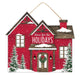 An enchanting 11.25"L x 9.75"H MDF house sign, named the Home/Holidays House (AP7174), captures the spirit of a cozy home with its red structure, brown and white snow-covered roofs, and inviting "Home For The Holidays" message. It features a festive wreath on the door, vibrant lights, delicate snowflakes, and is framed by two elegant evergreen trees on either side in green hues.